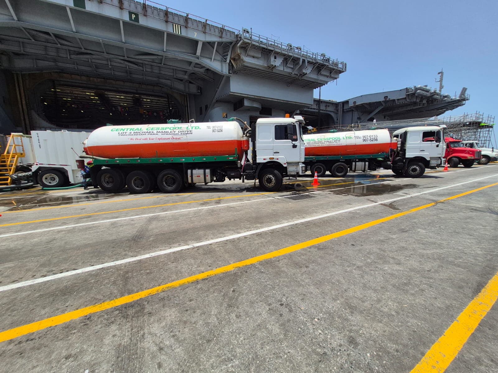 Two white and orange tanker trucks parked on a dock next to an aircraft carrier.