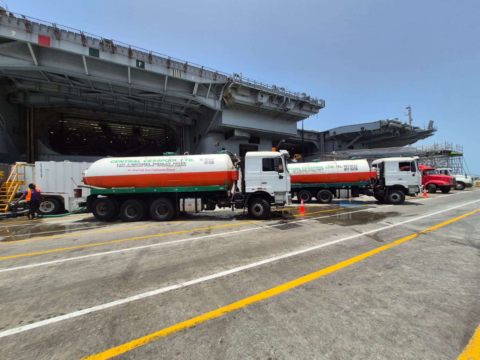 White and orange tanker trucks parked on a dock beside a massive grey aircraft carrier.