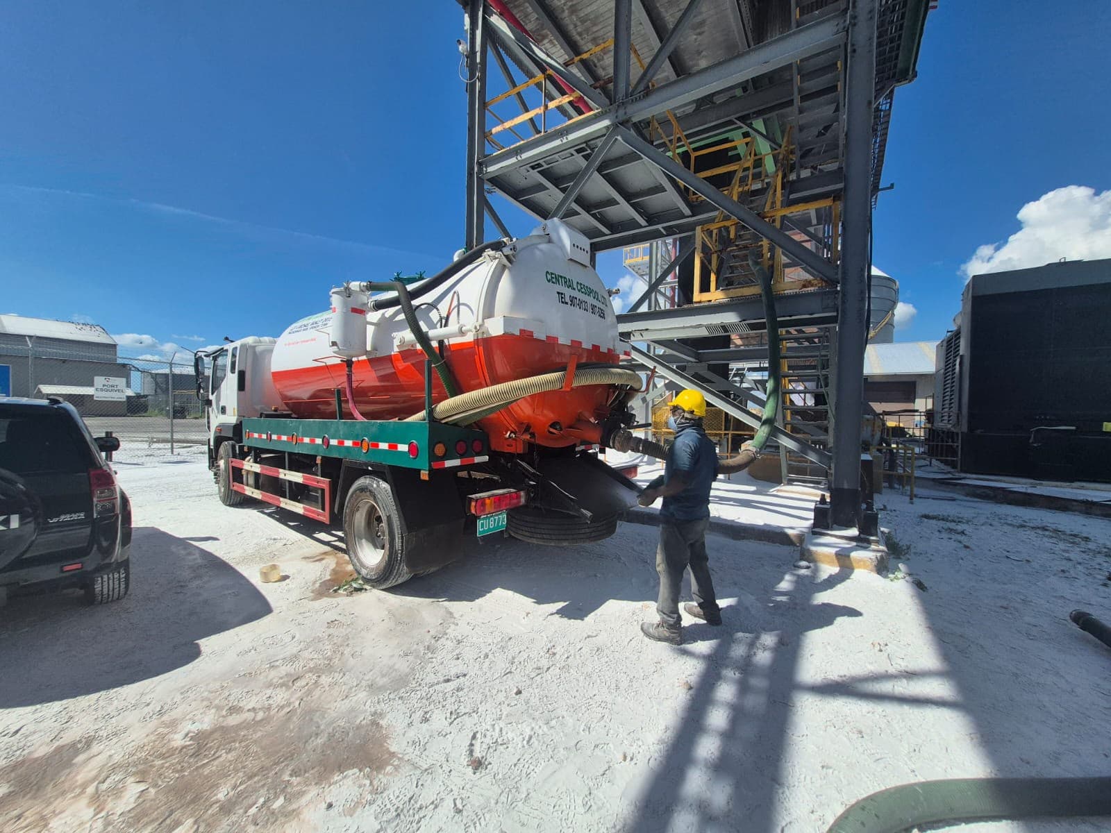 Worker in a yellow hard hat operating a vacuum truck at a white industrial site.