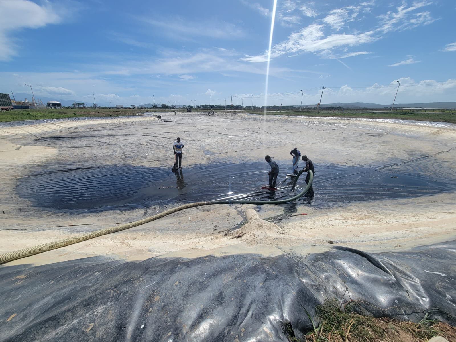 Workers cleaning dark sludge from a large lined basin using hoses and squeegees.