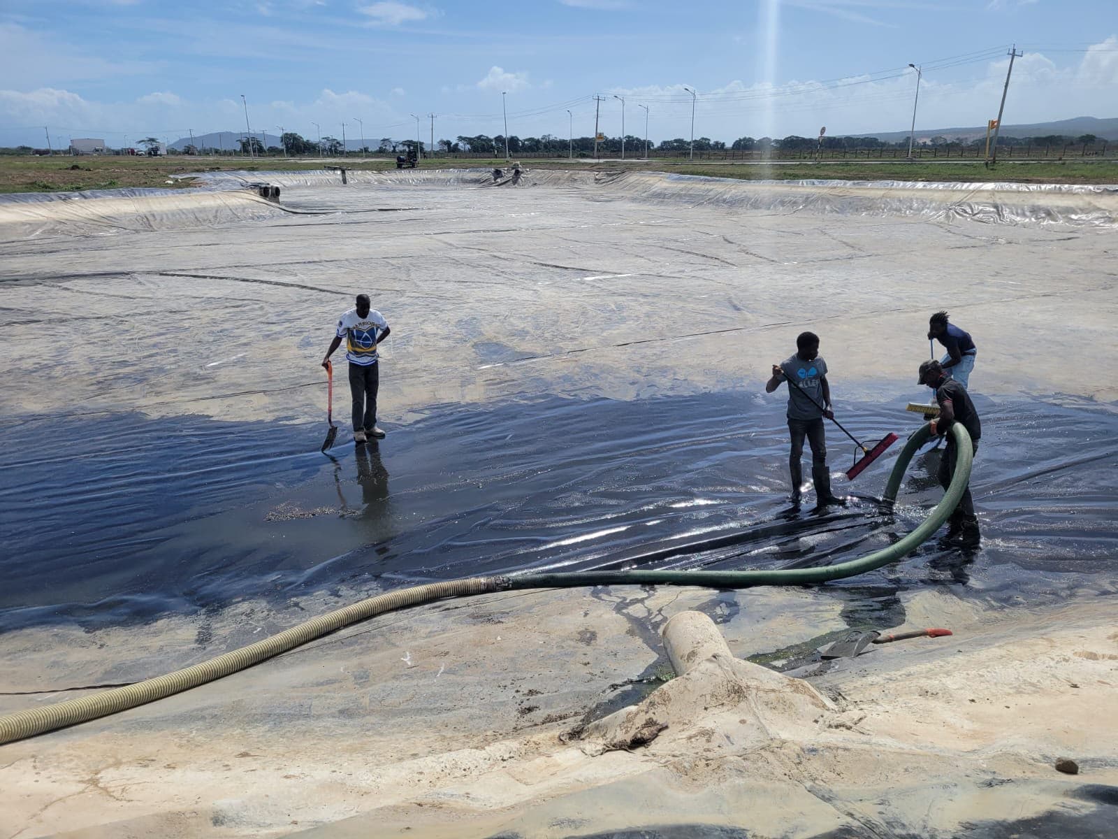 Workers cleaning a large lined wastewater basin using squeegees and a suction hose outdoors.