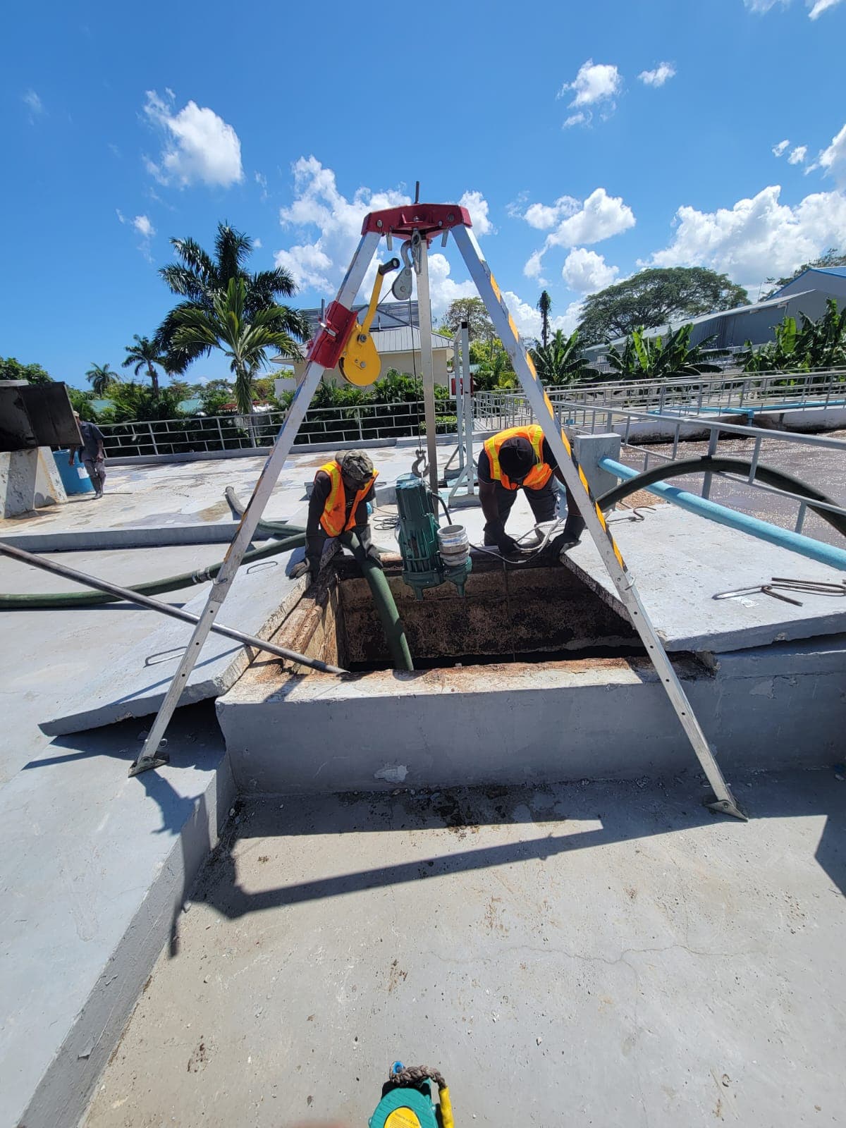 Workers in safety vests use a tripod hoist to lower equipment into a concrete opening.