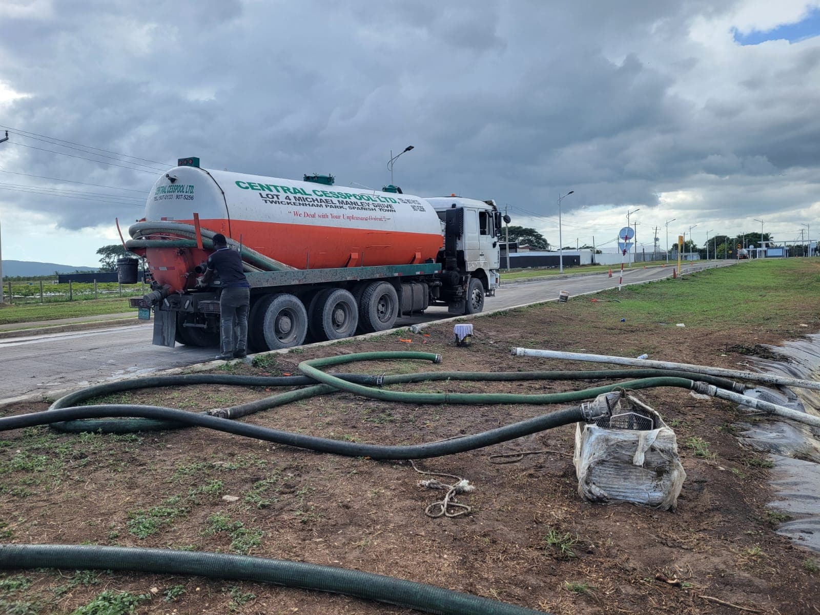 Worker operates a Central Cesspool tanker truck with long green hoses snaking across the dirt.