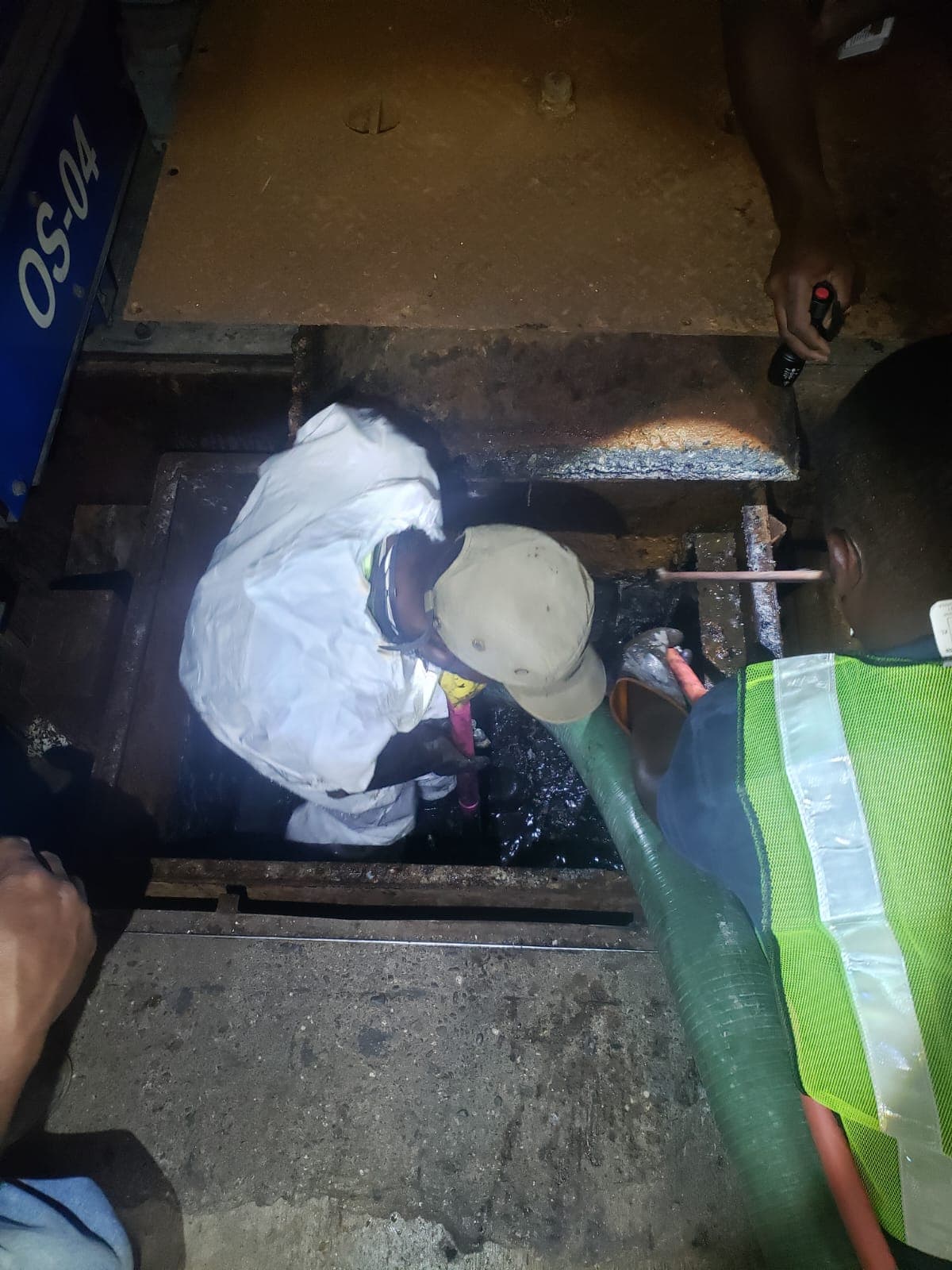Worker in protective gear cleans a dark manhole using a large green suction hose.